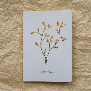 May include: A white notebook with a rounded top right corner featuring a watercolour illustration of thin green stems with orange, textured flower buds. The text 2027 Planner is printed in black ink at the bottom centre of the front cover.