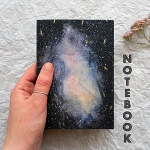 Op de afbeelding: Een notitieboek met een aquarel schilderij van een sterrenstelsel op de kaft. Het sterrenstelselontwerp heeft tinten blauw, roze en geel tegen een zwarte achtergrond, met kleine gouden steraccenten. Het woord "NOTEBOOK" is verticaal gedrukt.