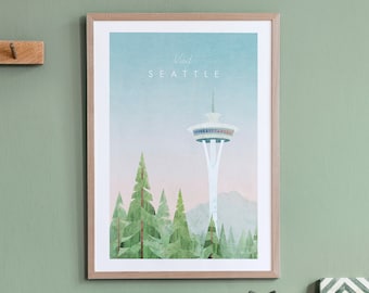 May include: A framed print with a minimalist design featuring the Space Needle in Seattle, Washington. The print has a light blue background with a pink gradient at the bottom. The text "Visit Seattle" is at the top of the print. The Space Needle is white with blue and red accents. There are green trees in the foreground.