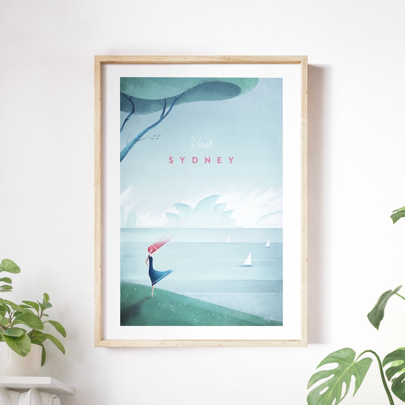 May include: Framed art print featuring a stylized illustration of Sydney. The artwork depicts a figure with flowing red hair and a blue dress, standing near the water. The text "Visit SYDNEY" is displayed in pink. The print is in a light wood frame.