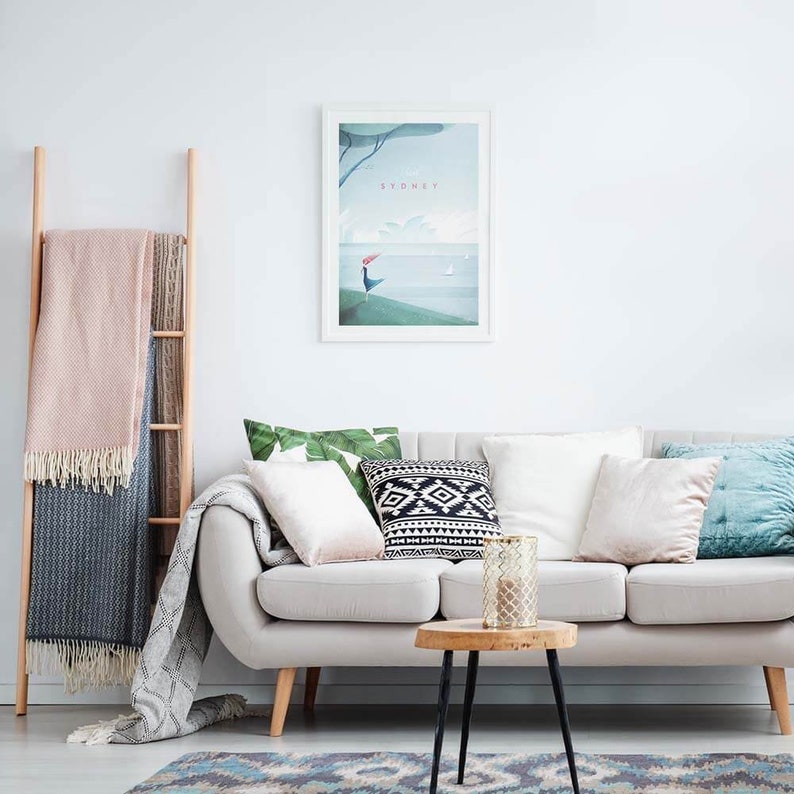 May include: A living room scene with a light gray sofa, pillows, and a wooden side table. A framed print with the word "SYDNEY" hangs on the wall. A wooden ladder holds blankets, and a patterned rug is on the floor.