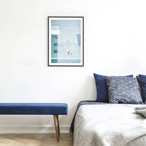 May include: A blue velvet bench with wooden legs sits in front of a bed with a white duvet and blue and grey patterned pillows. A framed poster with the text "Copenhagen" hangs on the wall above the bed.