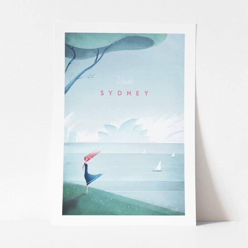 May include: A framed art print featuring a stylized illustration of Sydney, Australia. The artwork depicts a person standing near the water with the Sydney Opera House in the background. The text "Visit SYDNEY" is displayed in pink.