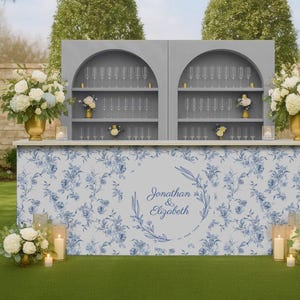 May include: A wedding bar with a blue floral design, featuring champagne flutes and floral arrangements. The bar has a gray backdrop with arched shelves. The text "Jonathan & Elizabeth" is displayed on the front.