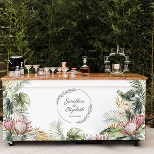 May include: A white bar cart with a wooden top and tropical floral design. The cart features a variety of glassware, bottles, and a cocktail shaker. The cart has the names "Jonathan & Elizabeth" and a date.