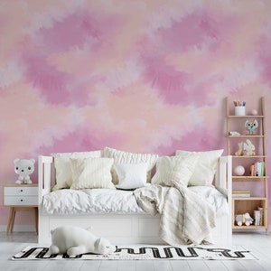 May include: A white day bed with a white blanket and pillows in a room with a pink and orange watercolour wall mural. A wooden shelf with books and toys is in the corner of the room.