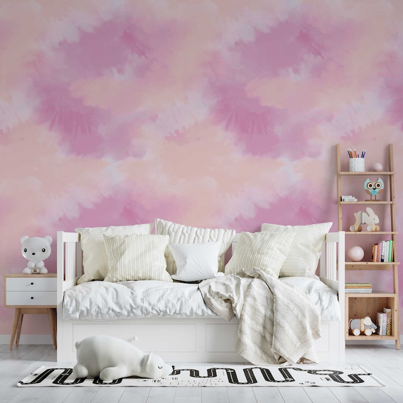 May include: A white day bed with a white blanket and pillows in a room with a pink and orange watercolour wall mural. A wooden shelf with books and toys is in the corner of the room.