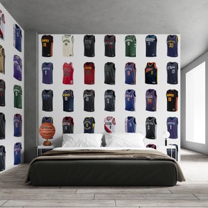 May include: A white wall with a basketball jersey collage featuring various NBA teams. The jerseys are arranged in rows and columns, with each jersey displaying a different team's logo and number. A basketball is on a stand in front of the wall, and a bed with a black headboard and a white bedspread is in the foreground.
