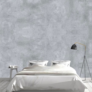 May include: A white bed with a white duvet and two white pillows sits in front of a gray concrete wall. A black lamp with a gold arm stands on a tripod base to the right of the bed.