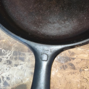May include: Close-up of a well-seasoned, black cast iron skillet. The handle is visible, with the number "10" embossed on it. The skillet's cooking surface shows signs of use, indicating it's ready for cooking.