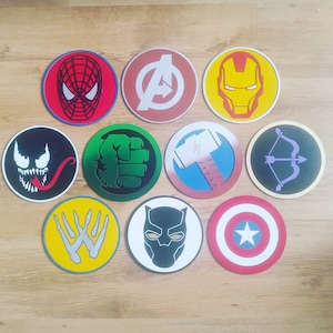 May include: A set of circular coasters displaying various superhero emblems. Designs include Spider-Man, the Avengers logo, Iron Man, Venom, Hulk's fist, Thor's hammer, Hawkeye's bow, a WWE logo, Black Panther, and Captain America's shield.