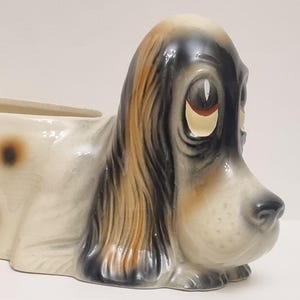 Op de afbeelding: Een keramische hondenplanter in liggende positie. De hond is voornamelijk crèmekleurig met bruine vlekken en zwart- en bruinaccenten op de kop en oren. De planter heeft een rechthoekige opening voor planten.