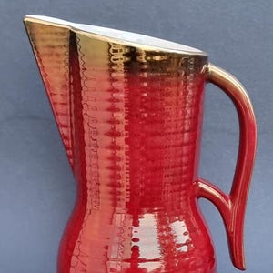 May include: A ceramic pitcher featuring a deep red, ribbed body with a glossy finish. The rim, handle, and base are accented with a metallic gold lustre. The pitcher has a tall, curved spout and a singular handle extending from the side.