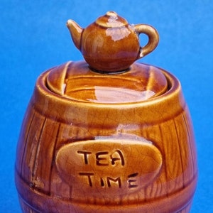 Op de afbeelding: Een bruine keramische theepot in de vorm van een vat met een deksel waarop een klein theepotje staat. De pot heeft de tekst "TEA TIME" aan de voorkant.
