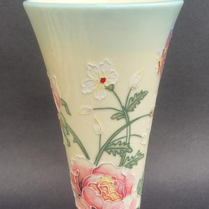 May include: A tall, flared ceramic vase with a cream-coloured base that transitions to a pale blue rim and a green base. The surface features raised, hand-painted floral motifs including a large pink rose and smaller white flowers with green leaves.