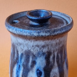 May include: A ceramic jar with a lid, featuring a mottled glaze in shades of brown, blue, and white. The jar has a rounded body and a slightly flared opening.