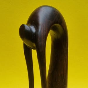 May include: A dark brown wooden sculpture with a smooth, polished finish. The abstract form features a curved, elongated shape with rounded elements. The sculpture is set against a bright yellow background, highlighting its form.