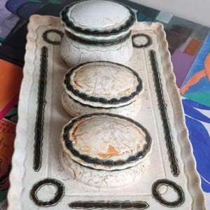 May include: A rectangular ceramic tray with scalloped edges featuring a cream-coloured crackle glaze and dark green decorative borders. Three matching round lidded ceramic containers with identical crackle glaze and dark green trim sit on top.