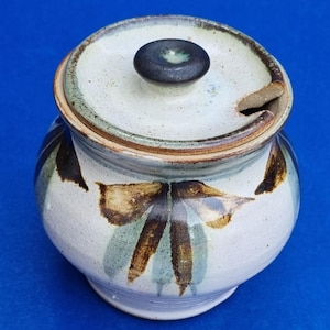 Peut inclure: Un pot en céramique blanche avec un couvercle et un motif de feuilles brunes et vertes. Le couvercle a un petit bouton sur le dessus.