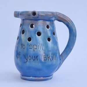 Peut inclure: Un pichet en céramique bleue avec une anse et un bec verseur, présentant de multiples trous circulaires sur la partie supérieure. La surface a une finition brillante avec des tons bleu foncé et marron. Le texte "not to spill of your skill." est écrit en noir.
