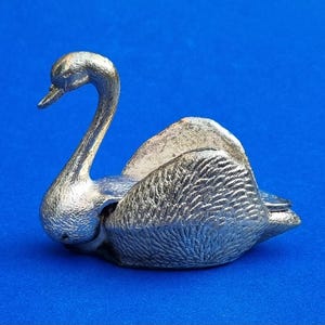 May include: A silver swan ornament with intricate feather detailing and a curved neck, set against a blue backdrop. This decorative piece could be used as a paperweight or a small trinket holder. The swan's design is elegant and detailed.