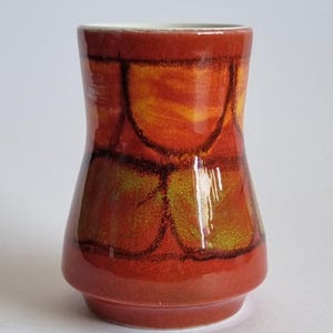 Peut inclure: Un vase en céramique avec une silhouette évasée et une finition brillante. L'extérieur présente un motif vibrant de formes arrondies orange et jaunes soulignées de brun foncé, créant un effet de grille sur une base rouge-orangé.