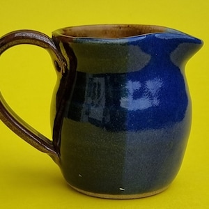 Op de afbeelding: Een kleine, blauwe en bruine keramische kan met een rond lichaam en een handvat. De kan is geglazuurd in een donkerblauwe kleur met een bruine rand en handvat.