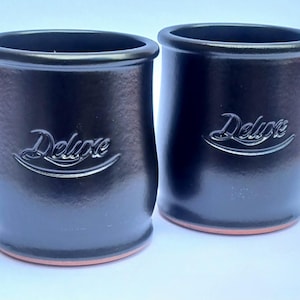 May include: Two black ceramic jars with a glossy finish. The word "Deluxe" is embossed on the front of each jar in a cursive font. The jars have a rounded shape with a slightly flared rim and a terracotta-coloured base.