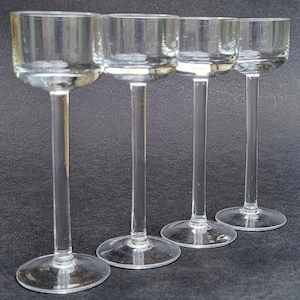 Op de afbeelding: Vier heldere glazen shotglazen met lange, dunne stelen. De glazen zijn in een rij gerangschikt, waarbij de stelen van links naar rechts steeds korter worden.
