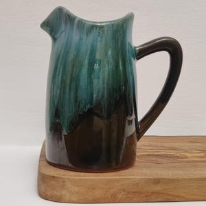 May include: A ceramic jug with a dark brown handle and base, fading into teal and green hues. The jug features a distinctive spout and is presented on a wooden chopping board.