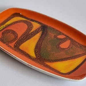 May include: A rectangular ceramic tray with rounded corners, featuring a vibrant orange border and a central abstract design in shades of yellow, dark brown, and muted green. The surface has a glossy finish with textured, dark brown linear accents.