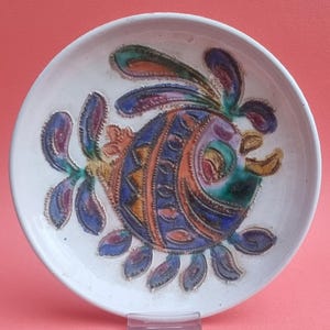 May include: A white ceramic plate with a colourful fish design. The fish features blue, orange, purple, and green details. The plate has a slightly raised edge and is likely handmade.