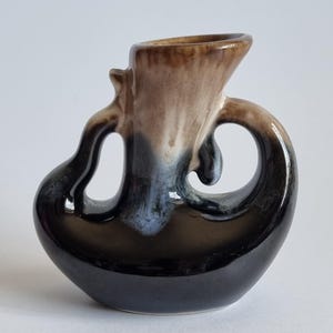 May include: A ceramic vase featuring a unique asymmetrical shape with two distinct handles. The glaze transitions from a dark black base to a lighter tan and cream colour at the top, creating a smooth, glossy finish with a curved, sculptural form.