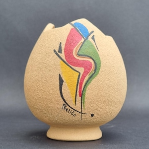 May include: A beige, textured ceramic vase with a rounded body and a pedestal base. The front features an abstract painted design in red, green, blue, and yellow, outlined in black. The name Bellon is written in black script near the bottom.