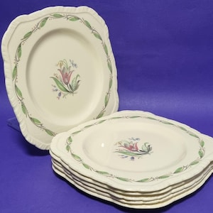 Op de afbeelding: Crème-witte keramische borden met een geschulpte rand en een bloemmotief. De borden hebben een groene en bruine bladrand en een centraal bloemmotief met roze, groene en blauwe accenten. Een stapel borden is zichtbaar.