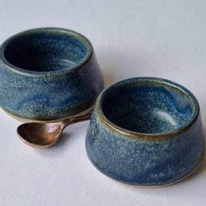 May include: Two small, round ceramic bowls with a mottled blue and brown glaze finish. The bowls have a wide base and a slightly tapered rim. A small, metallic copper-coloured spoon is positioned between the two bowls on the surface.