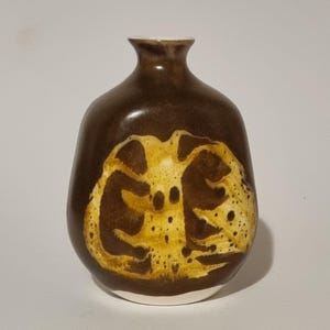 May include: A rounded ceramic vase with a dark brown glazed finish. The front features a prominent, abstract, light yellow and mustard-coloured organic pattern with speckled details. The vase has a narrow neck and a wide, bulbous body shape.