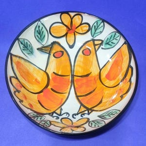 May include: A ceramic bowl with a black rim, featuring a hand-painted design of two orange birds facing each other. The birds have red eyes and are set against a white background with orange flowers and green leaves.