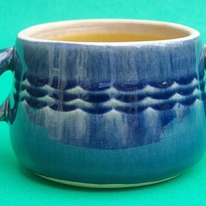 Op de afbeelding: Een blauwe keramische soepkom met twee handvatten. De kom heeft een golfachtig patroon in het midden en een crèmekleurige rand. De binnenkant van de kom lijkt een gele vloeistof te bevatten.