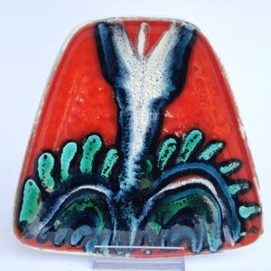 May include: A triangular ceramic dish with a glossy finish, featuring a vibrant red background. The centre displays a stylised, elongated white and dark blue floral motif, surrounded by smaller, textured turquoise and dark blue leaf-like shapes.
