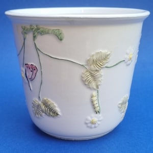 May include: White ceramic plant pot with embossed floral and insect designs. The pot features a strawberry plant motif with leaves, flowers, and a caterpillar. The design is in shades of green, pink, and cream.