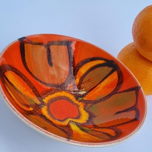 May include: A shallow, oval-shaped ceramic bowl featuring a vibrant orange and yellow floral pattern with bold, dark brown outlines. The interior surface is glossy, and the bowl is positioned next to two whole, textured orange citrus fruits.