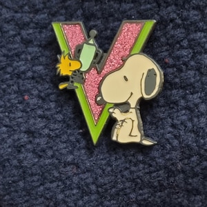 Peut inclure: Épingle en émail Snoopy et Woodstock. L'épingle présente un « V » scintillant rose avec un contour vert. Snoopy est assis et Woodstock est à côté d'un objet vert. L'épingle est sur un fond bleu foncé texturé.