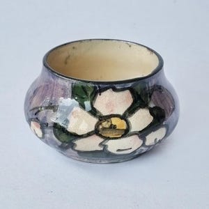 May include: A small, round ceramic bowl with a cream-coloured interior and a glossy exterior featuring a hand-painted floral design. The flower has white petals with dark outlines, green leaves, and a yellow centre, set against a purple wash.
