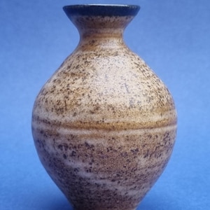 May include: A small, speckled ceramic vase with a rounded body and a narrow neck. The vase has a brown and tan colour scheme with a dark rim. The surface has a textured appearance, suggesting a handmade or artisanal quality.