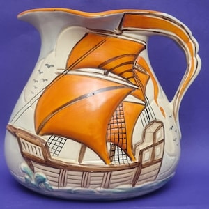 Op de afbeelding: Crèmekleurige keramische kan met een gedetailleerd oranje zeilschipontwerp. Het schip heeft oranje zeilen en bruine accenten, met een blauwe golf aan de basis. Decoratieve vogels zijn aan de zijkant geschilderd.