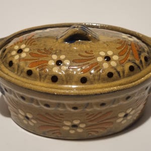 May include: An oval ceramic casserole dish with a matching lid, featuring a tan glazed finish. The exterior and lid are decorated with hand-painted floral patterns in cream, dark brown, and terracotta orange, with two side handles for lifting.