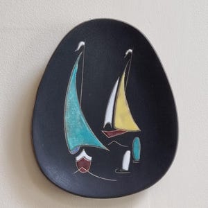 Peut inclure: Assiette décorative ovale noire avec un motif de voilier. L'assiette présente deux voiliers avec des voiles turquoise et jaunes, des accents blancs et une coque marron. L'assiette est fixée sur un mur beige clair.