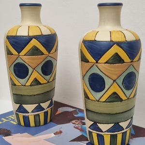 May include: Two decorative ceramic vases with cream-coloured bodies and geometric patterns in yellow, blue, green, and brown. The vases have a narrow neck and a blue rim. The design includes triangles, diamonds, and circles. The vases are displayed on a patterned surface.