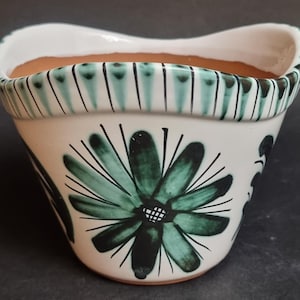 Peut inclure: Pot de fleurs en céramique blanche avec un motif floral vert. Le pot a un bord incurvé avec une bordure rayée verte et noire. Le motif floral est centré sur le devant du pot.